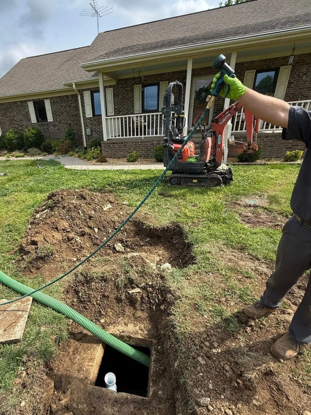 Green pump hose running across the yard into an opened septic tank lid in front of a brick house